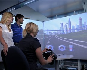 Studierende fahren gemeinsam an einer großen Leinwand virtuell Autorennen.