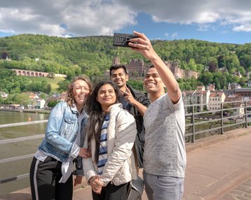 Studierende auf der Alten Brücke in Heidelberg, welche gerade ein Selfie mit Blick auf das Heidelberger Schloss schießen.