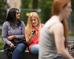 Eine Studentin blickt auf ihr Handy, die Freundin schaut in eine andere Richtung.