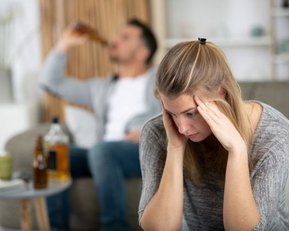 Eine junge Frau stützt den Kopf auf die Hände, während ein Mann im Hintergrund Alkohol trinkt.