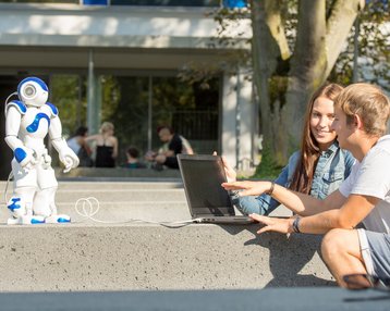 Studierende sitzen gemeinsam auf einer Mauer und steuern mittels Laptop einen Roboter