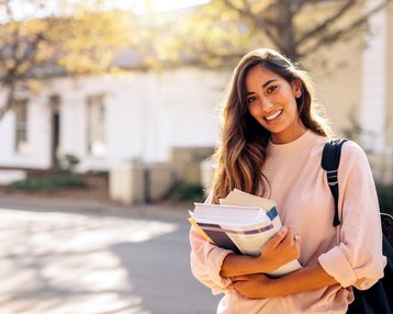 Studentin mit langen braunen Haaren und rosafarbenem Pullover steht mit Büchern in der Hand vor dem Gebäude